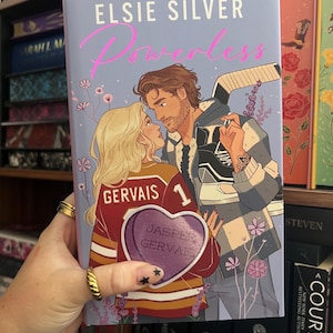 Può includere: Un libro con copertina rigida intitolato "Powerless" di Elsie Silver. La copertina presenta una coppia che si abbraccia, con una mazza da hockey e una maglia. La donna indossa una maglia con "Gervais 1" e una grafica a forma di cuore. Sfondo lilla chiaro con fiori.
