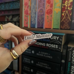 May include: A white sticker with the text "Little Monster" is held in front of a bookshelf filled with books. The sticker is rectangular and the text is in a simple sans-serif font. The books have colourful spines.