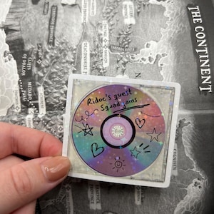 May include: A holographic sticker featuring a CD design with the text "Ridoc's quest Squad Jams." The sticker has colorful accents, including stars, hearts, and a smiling sun. The sticker is set against a map background.