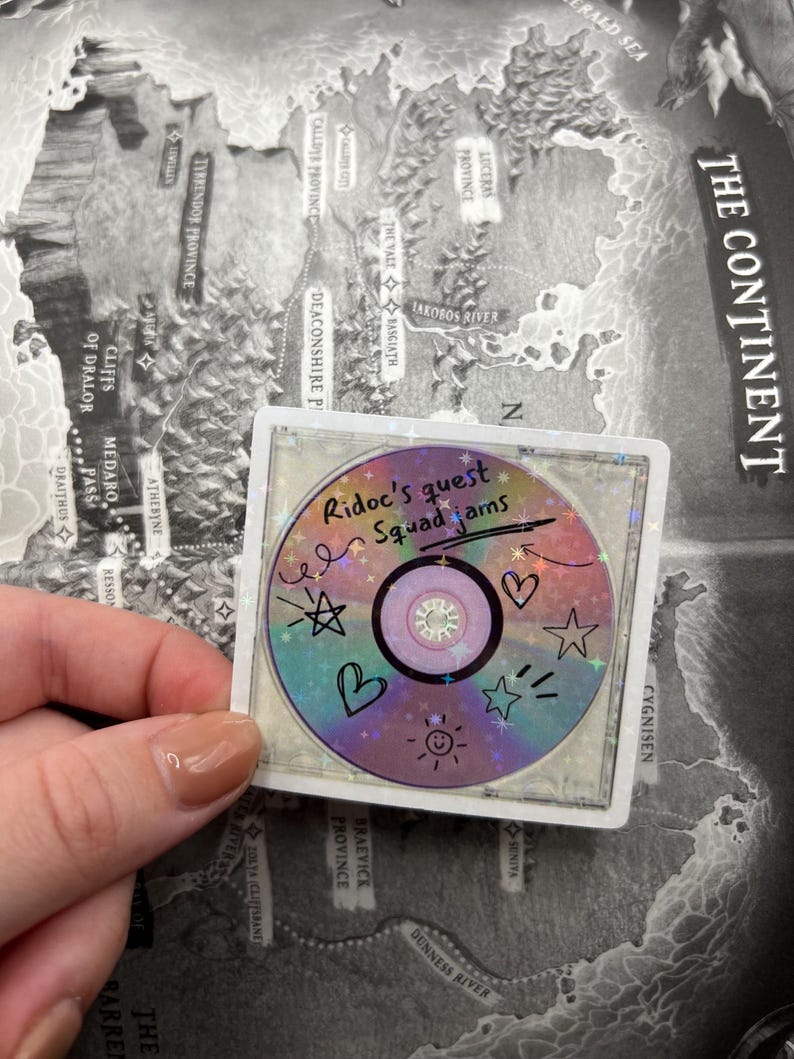 May include: A holographic sticker featuring a CD design with the text "Ridoc's quest Squad Jams." The sticker has colorful accents, including stars, hearts, and a smiling sun. The sticker is set against a map background.