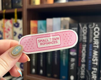 Peut inclure: Un autocollant rose en forme de pansement avec le texte "SMALL TOWN ROMANCES" en blanc. L'autocollant est tenu devant une bibliothèque remplie de livres. L'autocollant a une bordure pointillée.