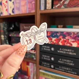 Puede incluir: Una pegatina con un dise&ntilde;o en forma de coraz&oacute;n y el texto "Johnny Kavanagh" y "13". La pegatina se sostiene frente a una estanter&iacute;a llena de libros. La pegatina tiene un dise&ntilde;o de lazo en la parte superior.