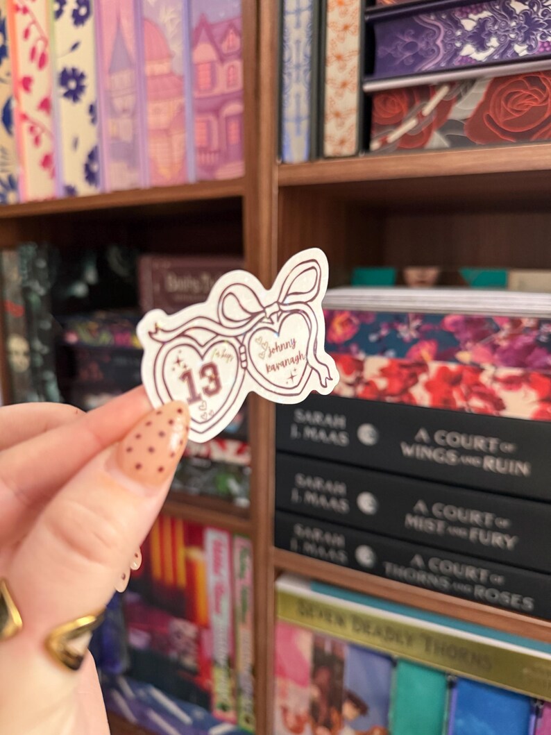 Puede incluir: Una pegatina con un dise&ntilde;o en forma de coraz&oacute;n y el texto "Johnny Kavanagh" y "13". La pegatina se sostiene frente a una estanter&iacute;a llena de libros. La pegatina tiene un dise&ntilde;o de lazo en la parte superior.