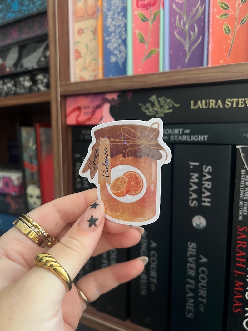 Op de afbeelding: Een sticker met een pot sinaasappelmarmelade met de tekst "Choose Violence". De pot is bruin met een etiket en een strik. De achtergrond toont een boekenplank met boeken.
