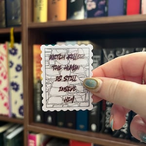 Könnte beinhalten: Ein weißer Aufkleber mit einem Briefmarkendesign, der den Text "Witch Killer The Human Is Still Inside Him" in handschriftlicher Form trägt. Der Aufkleber wird vor einem Bücherregal mit Büchern gehalten.