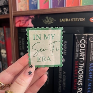Peut inclure: Un autocollant vert avec un design de timbre-poste, portant l'inscription "IN MY Sci-Fi ERA" dans une police manuscrite. L'autocollant est tenu devant une étagère remplie de livres.