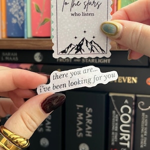 May include: Two white stickers with black text are held in front of a bookshelf. One sticker reads "to the stars who listen" with a mountain illustration. The other says "there you are... i've been looking for you."