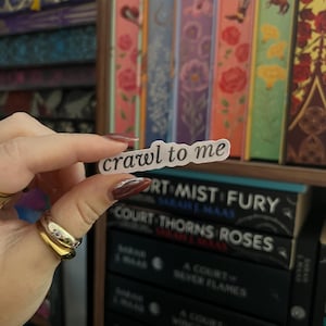 Può includere: Un adesivo trasparente con la scritta "crawl to me" tenuto davanti a una libreria. Lo sfondo presenta una collezione di libri con dorsi colorati. I libri sono neri con testo bianco.