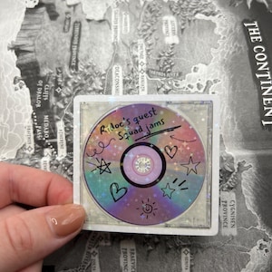 May include: A holographic sticker featuring a colorful CD design with the text "Ridoc's quest squad jams." The CD is decorated with doodles of stars, hearts, and a smiling sun. The sticker is held over a map.