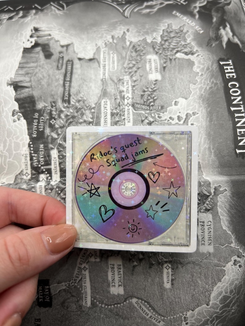 May include: A holographic sticker featuring a colorful CD design with the text "Ridoc's quest squad jams." The CD is decorated with doodles of stars, hearts, and a smiling sun. The sticker is held over a map.