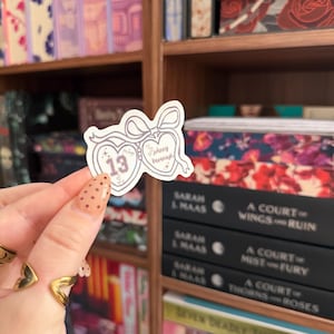 Puede incluir: Una pegatina en forma de coraz&oacute;n con el n&uacute;mero 13 y el nombre "Johnny Baranagh". La pegatina es de color lila claro con un borde blanco y un lazo. El fondo muestra estanter&iacute;as llenas de libros.