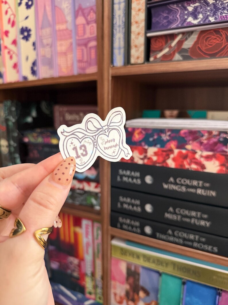 Puede incluir: Una pegatina en forma de coraz&oacute;n con el n&uacute;mero 13 y el nombre "Johnny Baranagh". La pegatina es de color lila claro con un borde blanco y un lazo. El fondo muestra estanter&iacute;as llenas de libros.