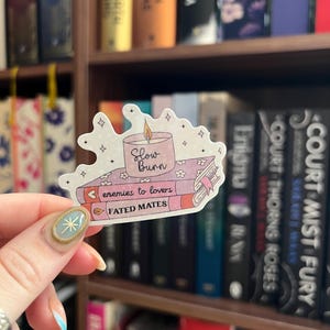 May include: A sticker featuring a candle with the text "Slow Burn" on top of three books. The books are labeled "enemies to lovers" and "FATED MATES". The sticker has a watercolor style with pink, orange, and white colors.