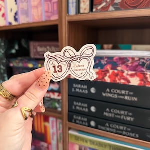 Puede incluir: Una pegatina con un dise&ntilde;o en forma de coraz&oacute;n, con el n&uacute;mero 13 y el nombre "Johnny Kavanagh" en una fuente manuscrita. La pegatina se sostiene frente a una estanter&iacute;a llena de libros.