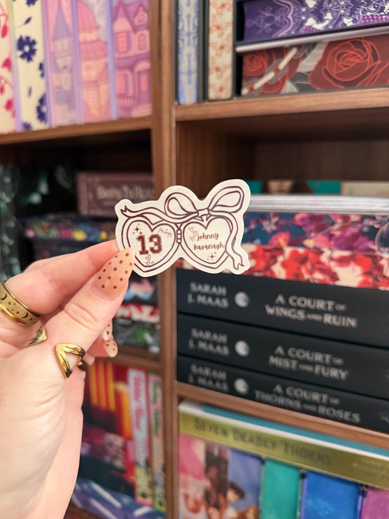Puede incluir: Una pegatina con un dise&ntilde;o en forma de coraz&oacute;n, con el n&uacute;mero 13 y el nombre "Johnny Kavanagh" en una fuente manuscrita. La pegatina se sostiene frente a una estanter&iacute;a llena de libros.