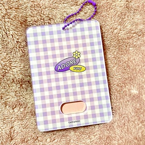 May include: A rectangular card holder with a purple and white gingham pattern. The holder features the text "ADORE YOU" with a flower graphic. It has a small pink section and a purple beaded chain.