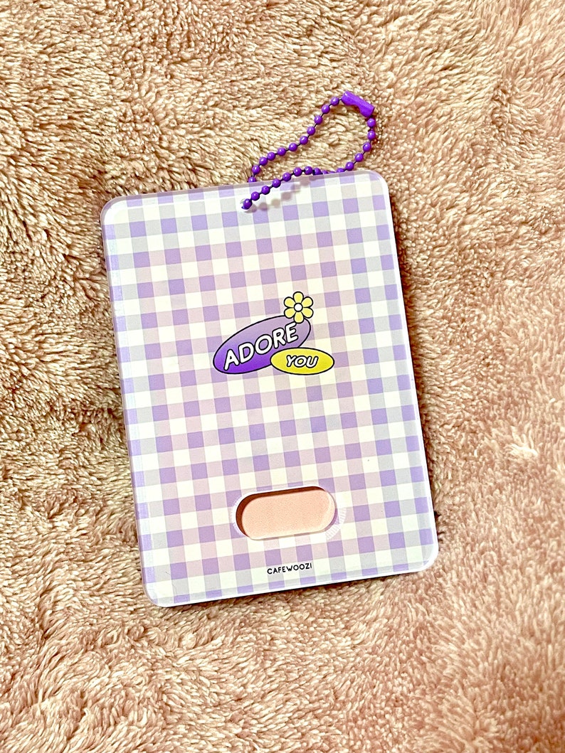 May include: A rectangular card holder with a purple and white gingham pattern. The holder features the text "ADORE YOU" with a flower graphic. It has a small pink section and a purple beaded chain.