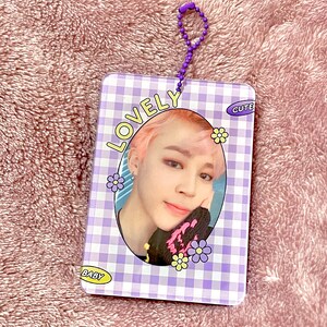 May include: A rectangular, purple and white checkered card with a photo of a person with pink hair. The card has the words "LOVELY", "CUTE", and "BABY" printed on it, along with flower graphics. A purple beaded chain is attached to the top.