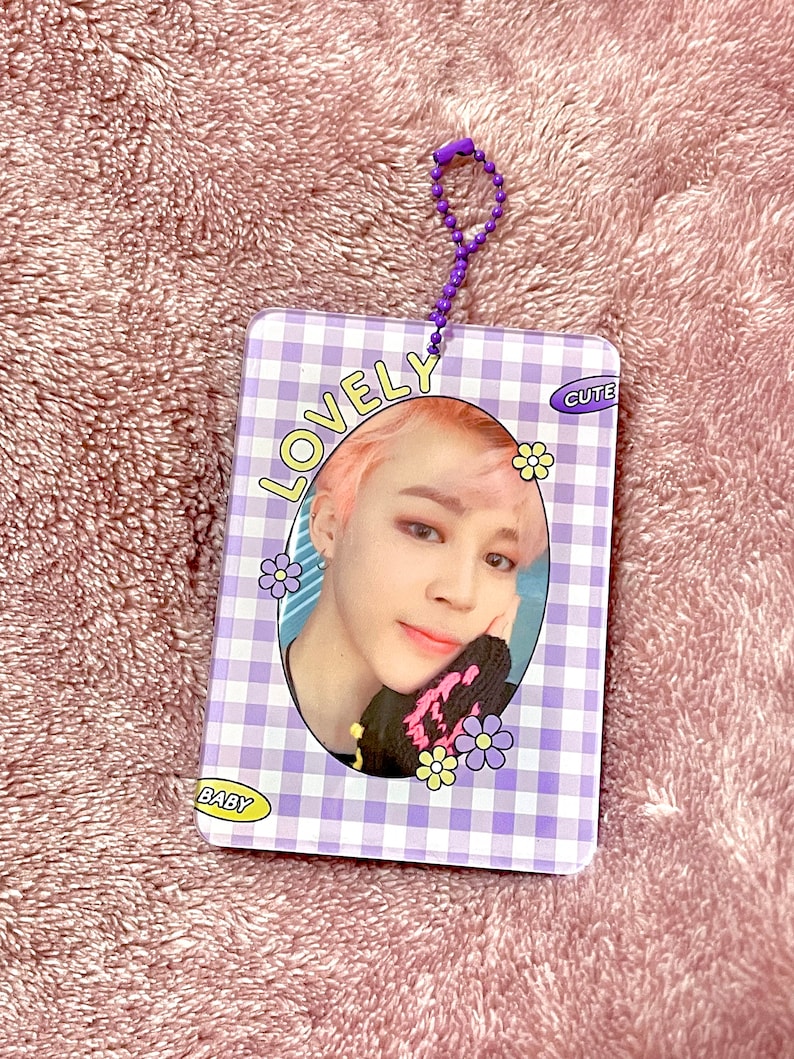 May include: A rectangular, purple and white checkered card with a photo of a person with pink hair. The card has the words "LOVELY", "CUTE", and "BABY" printed on it, along with flower graphics. A purple beaded chain is attached to the top.