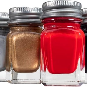 May include: A collection of small glass jars filled with various colors of paint, including yellow, white, silver, bronze, red, black, green, blue, and clear. Each jar has a silver metal lid. A paintbrush is visible on the left.