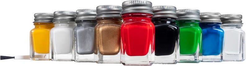 May include: A collection of small glass jars filled with various colors of paint, including yellow, white, silver, bronze, red, black, green, blue, and clear. Each jar has a silver metal lid. A paintbrush is visible on the left.