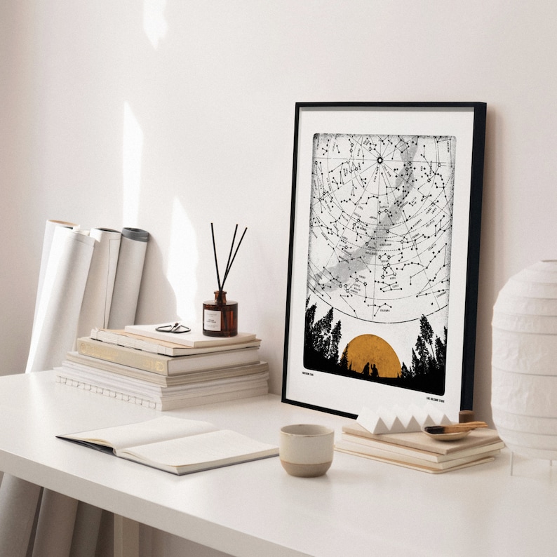 Puede incluir: Una impresi&oacute;n enmarcada en negro con fondo blanco y una ilustraci&oacute;n en blanco y negro de un mapa de constelaciones con un sol dorado. La impresi&oacute;n est&aacute; colgada en una pared blanca sobre una pila de libros y una mesa blanca.