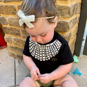 May include: A baby wearing a black t-shirt with a white collar design and the text "RBG" on the bottom. The baby is wearing a white bow in their hair and is sitting on a concrete surface.