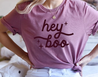 May include: A mauve t-shirt with the text "hey boo" in a dark red color. The shirt has a relaxed fit and is worn by a person with blonde hair.