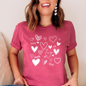 Pode incluir: Uma camiseta rosa com uma estampa gráfica branca de vários corações, alguns com palavras como "abraços", "mwah", "amor" e "xo".