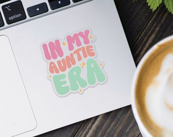 May include: A sticker on a laptop with the text "IN MY AUNTIE ERA" in pink, peach, and green bubble letters. The sticker has a white border and small star accents. The laptop is on a wooden surface, with a cup of coffee in the background.