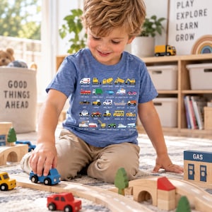 May include: A heather blue short-sleeve t-shirt featuring a graphic print of various vehicles including an ambulance, fire truck, and school bus, each labeled with its name in small white text arranged in a grid pattern on the front.