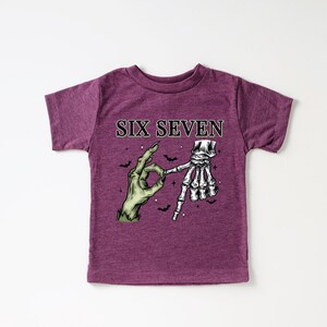 Puede incluir: Camiseta granate con el texto "SIX SEVEN" sobre un gráfico de una mano de zombi verde haciendo el signo "OK" y una mano de esqueleto. Pequeños murciélagos negros están dispersos.