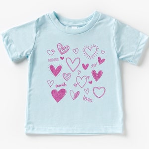 Pode incluir: Camiseta azul claro com um design de corações rosa. A camiseta tem as palavras "hugs", "mwah", "love" e "xo" impressas nela.