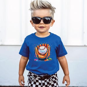 May include: A young child wearing a royal blue t-shirt with a baseball-themed graphic. The graphic features a baseball inside a baseball glove, with colorful paint drips and confetti. The child is also wearing black sunglasses.