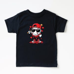 Op de afbeelding: Zwart T-shirt met een cartoon honkbalpersonage met een zonnebril en een rode pet, met een knuppel. De honkbal heeft rode accenten en is omgeven door harten. Het personage draagt rode sneakers.