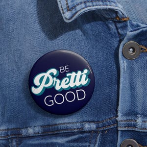 May include: A navy blue button with the words "BE Pretti GOOD" in white and teal script. The button is round and is set against a denim jacket.