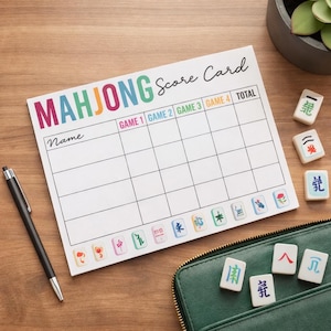 May include: A white Mahjong score card with the word "MAHJONG" in colorful letters. The card has spaces for game scores and the word "Name". A black pen and several Mahjong tiles are next to the card.