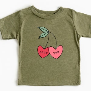 Pode incluir: Uma camiseta verde de manga curta com um gráfico de duas cerejas vermelhas. As cerejas têm as palavras "LOVE" e "YOU" escritas nelas.