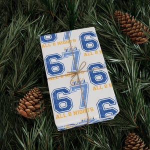 May include: A wrapped gift with a white background and a repeating pattern of the numbers 6 and 7 in blue, with the text "ALL 8 NIGHTS". The gift is tied with twine and rests on a bed of green pine needles and pine cones.