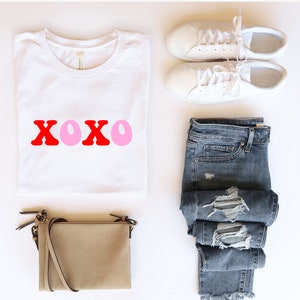 Pode incluir: Camiseta branca com a palavra "XOXO" em letras vermelhas e rosa. A camiseta é combinada com jeans azul desbotado, uma bolsa transversal bege e tênis brancos.