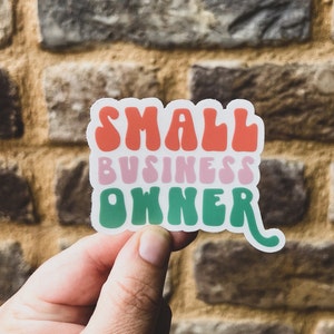 May include: A white sticker with the text "SMALL BUSINESS OWNER" in a retro font. The words are in different colors: "SMALL" is red, "BUSINESS" is pink, and "OWNER" is green.