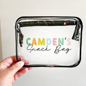 May include: Clear plastic snack bag with a black zipper and black trim. The bag has the text "CAMDEN'S Snack Bag" printed on it in colourful letters.