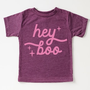 Puede incluir: Una camiseta de manga corta en color ciruela jaspeado con el texto "hey boo" impreso en una fuente cursiva de color rosa claro. El diseño incluye pequeños acentos de estrellas de cuatro puntas sobre y bajo el texto, centrado al frente.