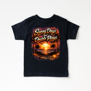 May include: A navy blue t-shirt featuring a vibrant graphic of a sunset over the ocean, framed by palm trees. The text "Sunny Days and Double Plays" is displayed in a stylized font. The design incorporates orange, yellow, and purple hues.