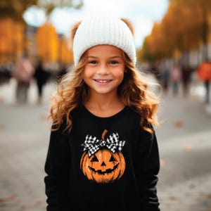 Op de afbeelding: Een jong persoon draagt een zwart sweatshirt met een pompoenontwerp en een witte gebreide muts met bruine pompons. De pompoen heeft een zwart-wit geruite strik. De achtergrond is wazig met herfstkleuren.