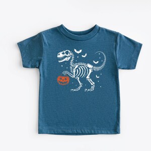 May include: A blue t-shirt with a white dinosaur skeleton holding an orange pumpkin. The design includes white bats and stars, ideal for Halloween. The shirt is made of a soft material.