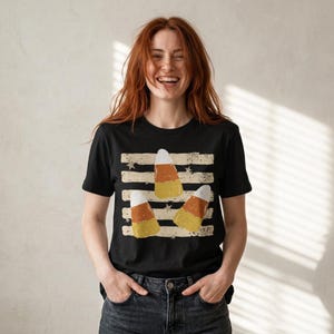 May include: A black short-sleeve t-shirt featuring a graphic print of three candy corn pieces arranged over horizontal distressed stripes and small star shapes. The design uses a weathered, vintage-style texture in shades of orange, yellow, and white.