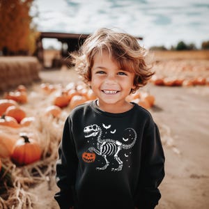 May include: A black sweatshirt with a white dinosaur skeleton design holding a pumpkin. The design includes bats and stars. The background shows a pumpkin patch and a child smiling.