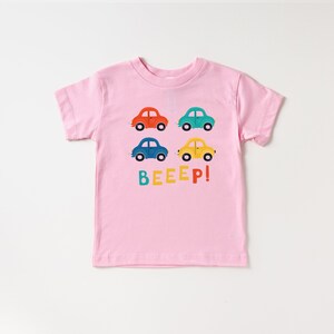 May include: Pink short-sleeved t-shirt with a graphic of four colourful toy cars and the text "BEEEP!" printed on the front.