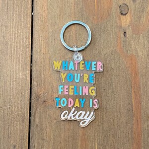 May include: A clear acrylic keychain with the text "Whatever you're feeling today is okay" in colorful letters. The keychain is attached to a silver metal ring.
